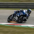 A MotoGP rider in a blue and white custom livery leans at an extreme angle through a corner, with his knee slider sparking on the track's kerbing.