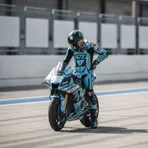 A MotoGP rider on a light blue and red motorcycle leans at an extreme angle while cornering on a race circuit.