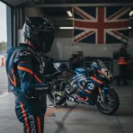 A veteran British rider stands in his pit garage, with his race bike and a Union Jack flag behind him.