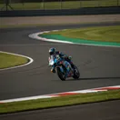A MotoGP rider leans their motorcycle at high speed through a fast, flowing corner at the Silverstone circuit.